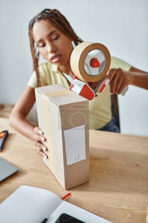Foto de Se centran en la caja de cartón con la mujer afroamericana borrosa usando cinta adhesiva, concepto de entrega - Imagen libre de derechos
