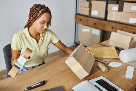 fröhliche junge afrikanisch-amerikanische Verkäuferin hält Klebeband in der Hand und schaut auf Karton, Lieferung