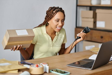 cheerful african american female merchant looking at laptop while scanning box, delivery concept