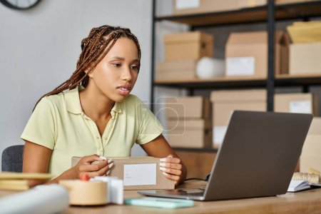 Foto de Atractiva mujer afroamericana comerciante mirando su computadora portátil mientras trabaja duro, entrega - Imagen libre de derechos