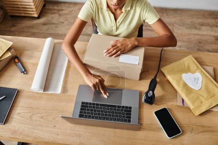 Photo for Cropped view of young african american female retailer packing box and working on laptop, delivery - Royalty Free Image