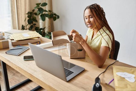 jolly young african american female seller packing box and looking at laptop, delivery concept