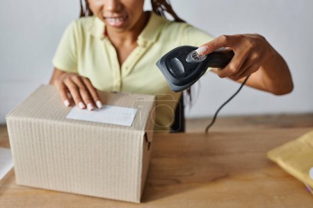 Téléchargez les photos : Vue recadrée avec mise au point sur la boîte en carton avec flou vendeur femme afro-américaine numérisation - en image libre de droit