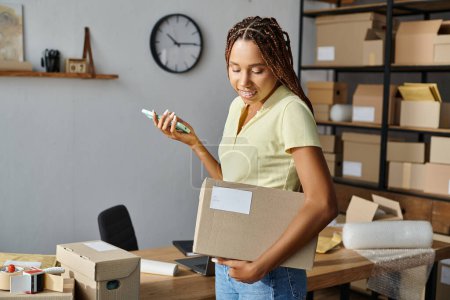 attractive african american female seller holding phone and looking at cardboard box, delivery
