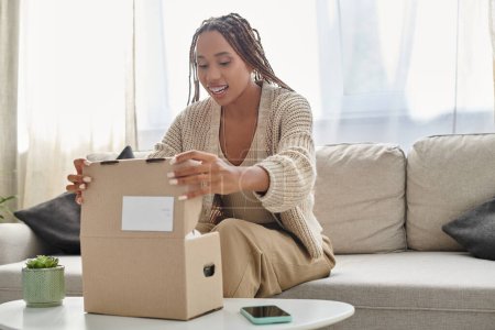 fröhliche junge afrikanisch-amerikanische Frau in kuscheliger Homewear sitzt auf dem Sofa und öffnet ihr Paket