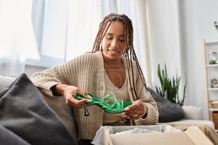 Téléchargez les photos : Joyeuse belle femme afro-américaine en tenue de maison assis et regardant ses nouvelles chaussures vertes - en image libre de droit