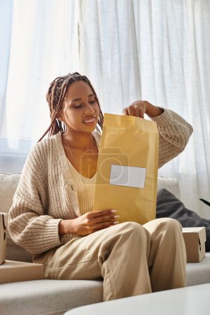 fröhliche junge afrikanisch-amerikanische Frau mit Zahnspange auf Couch sitzend und Postpaket öffnend
