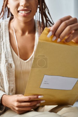 Téléchargez les photos : Vue recadrée avec accent sur le paquet postal dans les mains d'une femme afro-américaine joyeuse et floue avec des bretelles - en image libre de droit