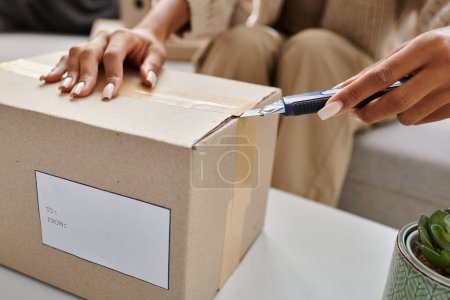 cropped view of young african american woman opening box with stationery knife next to plan on table