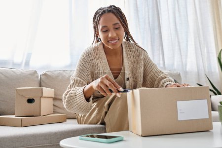 jolly african american woman in casual attire using stationery knife to open cardboard box