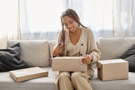 Téléchargez les photos : Belle femme afro-américaine joyeuse en tenue de maison assise sur le canapé et regardant la boîte en carton - en image libre de droit