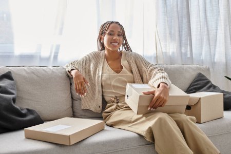 freudige junge afrikanisch-amerikanische Frau in kuscheliger Kleidung sitzt auf einem Sofa mit Schachteln und lächelt in die Kamera