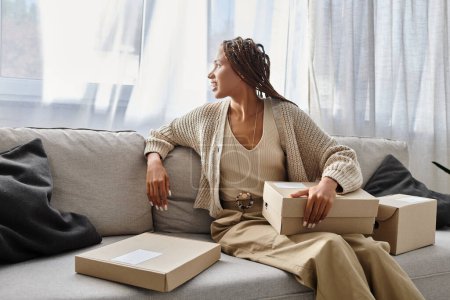 Téléchargez les photos : Femme afro-américaine gaie en vêtements de maison assis sur le canapé avec des boîtes en carton et regardant loin - en image libre de droit