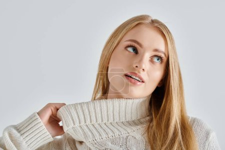 charming and dreamy woman stretching collar of white knitted sweater and looking away on grey