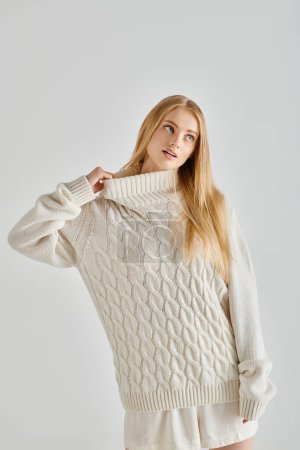 mujer rubia pensativa estiramiento cuello de suéter de invierno caliente mientras mira hacia otro lado en el fondo gris