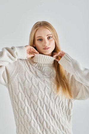 sensual dreamy woman with blonde hair touching collar of warm sweater and looking at camera on grey