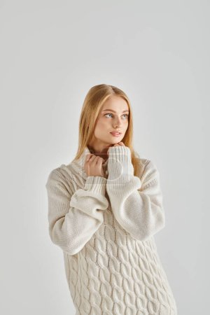 contemplative blonde woman in white warm sweater looking at camera on grey, winter emotions