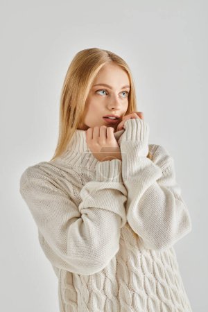 modelo femenino hipnotizante y soñador en suéter de invierno blanco mirando hacia otro lado mientras está de pie en gris