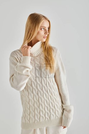 jeune femme rêveuse aux cheveux blonds posant en pull tricoté doux et regardant loin sur gris, hiver