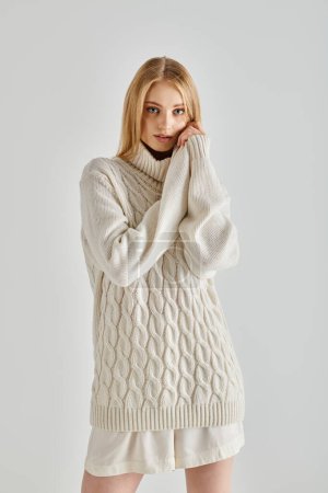 mesmerizing and contemplative woman in white knitted sweater looking at camera on grey, cozy winter