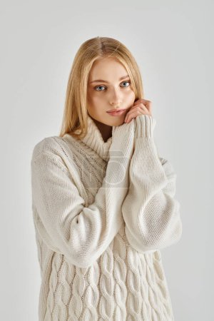 romantische und verträumte Frau mit blonden Haaren posiert in weichem warmen Pullover auf grauen, winterlichen Gefühlen