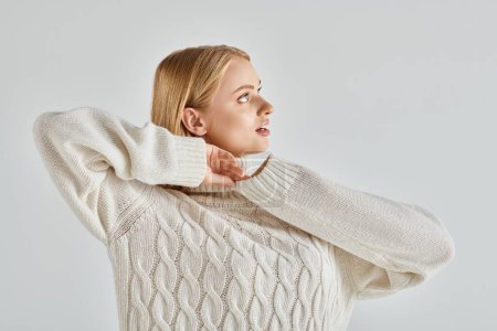 attractive and dreamy woman in warm sweater standing and looking away on grey, winter style