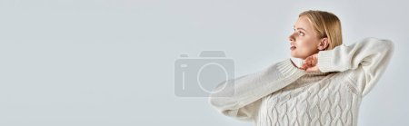 young and dreamy woman in warm and cozy winter sweater looking away on grey, horizontal banner