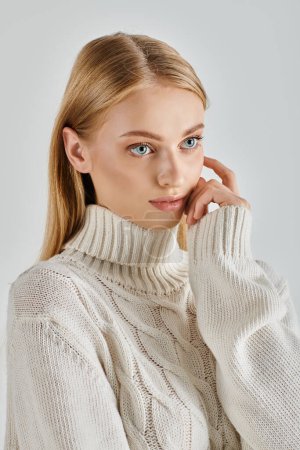 attractive and dreamy woman in white warm sweater touching face and looking away on grey backdrop
