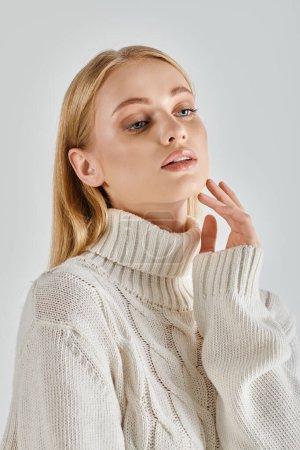 mujer rubia soñadora con pelo rubio usando suéter blanco acogedor sobre fondo gris, encanto de invierno