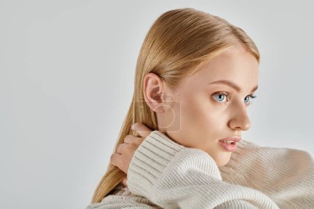 mesmerizing woman in white knitted sweater and natural makeup touching blonde hair on grey, portrait