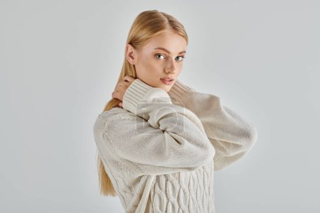 seductora mujer rubia en suéter de punto blanco abrazando su propio cuello y mirando a la cámara en gris