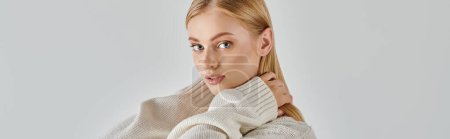 young blonde woman in soft knitted sweater embracing own neck and looking at camera on grey, banner