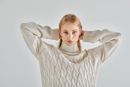 charming blonde woman in warm sweater holding hands behind head and looking at camera on grey