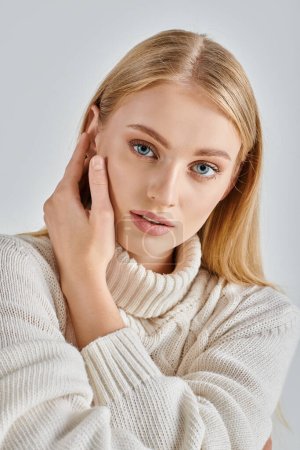 portrait of sensual blonde woman in natural makeup and white winter sweater touching face on grey