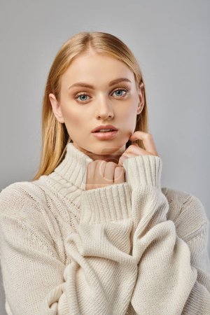 Nachdenkliche Frau mit blonden Haaren und natürlichem Make-up im weißen Winterpullover blickt in die Kamera auf grau