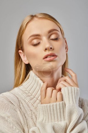 sensual blonde woman in white delicate sweater and natural makeup with closed eyes on grey, portrait