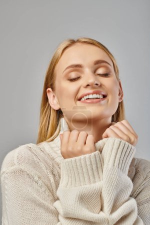 delighted blonde woman in warm knitted sweater smiling with closed eyes on grey, winter fashion