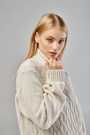 alluring woman with blonde hair and natural makeup wearing white sweater looking at camera on grey