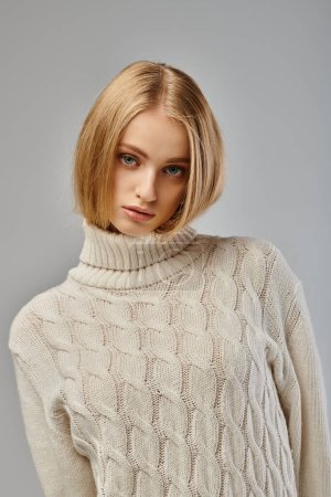 confident and charming blonde woman in white knitted sweater posing on grey, cold season clothing