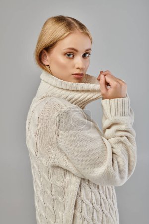 Téléchargez les photos : Jeune femme blonde en pull tricoté chaud avec col montant regardant la caméra sur gris, tendance hiver - en image libre de droit