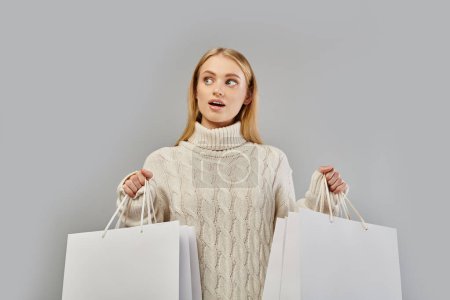Téléchargez les photos : Femme blonde étonnante en pull chaud blanc tenant des sacs à provisions blancs sur gris, les ventes d'hiver - en image libre de droit