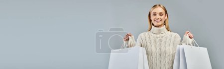 fröhliche blonde Frau im warmen Strickpullover mit weißen Einkaufstaschen auf grauem, waagerechtem Banner