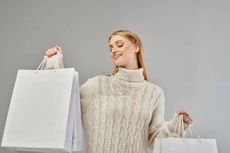 fröhliche blonde Frau im Strickwinterpullover posiert mit weißen Einkaufstaschen auf grau, Winterschlussverkauf