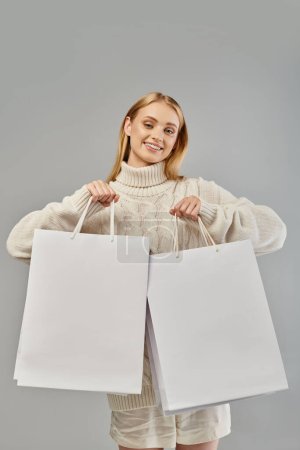 Téléchargez les photos : Joyeuse femme blonde en pull tricoté blanc montrant des sacs à provisions sur gris, ventes saisonnières - en image libre de droit