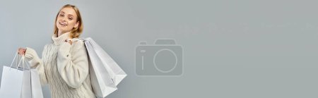 happy blonde woman in warm knitted sweater posing with white shopping bags on grey, banner