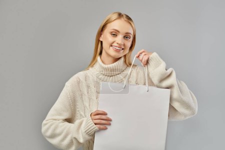 Frohe blonde Frau im warmen Strickpullover mit weißer Einkaufstasche auf grau, Winterschlussverkauf