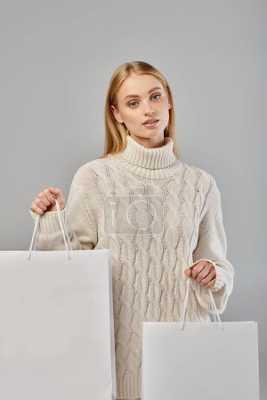 Charmante blonde Frau in weichem warmen Pullover mit weißen Einkaufstaschen auf grau, Winterschlussverkauf