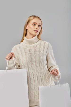Faszinierende blonde Frau in kuscheligem Strickpullover mit weißen Einkaufstaschen, die wegschauen auf grau