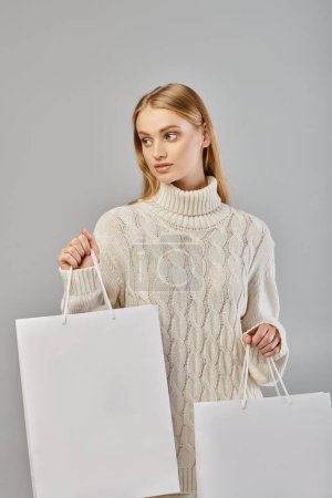 young blonde woman in white knitted sweater holding shopping bags and looking away on grey
