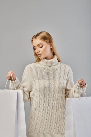 attraktive blonde Frau in warmem, stylischem Pullover mit weißen Einkaufstaschen auf grauem, saisonalem Einkauf